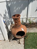 Full view of the terracotta chimenea on metal stand placed outdoors near a white fence and garden area. Visible wear and dirt on the surface.