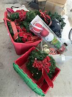 Red and green plastic bins filled with decorated green wreaths featuring large red bows and assorted ornaments, plus a clear bin of Christmas hanging ornaments in various colors.
