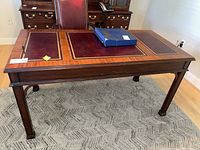 Front and side view of vintage Henckel Moore office desk showing leather inset top with gold tooling, three drawers with brass handles, and tapered legs, placed on a patterned rug.