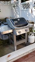 Weber Spirit II propane BBQ grill with black lid, temperature gauge, and two control knobs. Side tables and propane tank visible.
