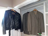Photo showing three jackets hanging on hangers: one black leather motorcycle jacket and two men's suit jackets (one beige, one patterned).