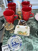 Four red Crate & Barrel mugs with red polka dots, blue and white Delft-style ashtrays, vintage brass cocktail skewers in a holder, miniature Wade pitchers, and Bavarian souvenir plate on green surface.