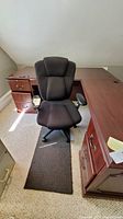 Black swivel office chair with padded back and seat, placed next to a wooden desk with a small dark-colored rug underneath.