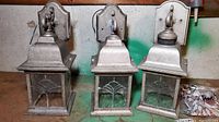 Three outdoor lantern light fixtures aligned, showing weathered metal finish and decorative glass with geometric pattern.