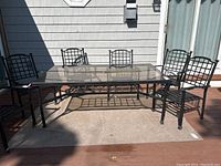 Patio table shown on outdoor deck with shadow cast from sunlight. Black metal frame with lattice design and clear glass top visible.