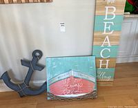 Photo showing three items: a wooden anchor painted dark blue-gray with rope hanging, a beach-themed print of a boat with text 'Let Your Dreams Set Sail' accented by driftwood, and a tall wooden plank wall hanging painted in teal and natural wood with 'The Beach is Calling' text.