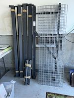 Metal shelving components leaning against wall in garage, showing black vertical supports and silver wire grid shelves.