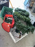 Multiple artificial green Christmas wreaths arranged on the floor next to a plastic bin and a red collapsible bag filled with string lights.