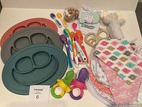 Overview of baby feeding and teething items including silicone plates, spoons, fruit feeders, bibs, and various teethers laid out on a surface.