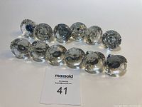 Twelve clear faux crystal drawer knobs arranged in two rows on a white surface with a MaxSold label.