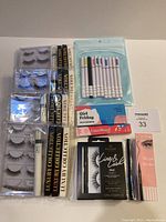 Four sets of false eyelashes, adhesive liners in black and white, colorful adhesive liners in pens, and a pink makeup remover pen arranged on a flat surface.