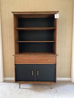 Full front view of the mid-century modern shelf unit showing three open shelves, one drawer with brass handles, and two cabinet doors with black vinyl panels and brass handles.