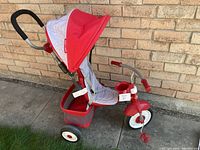 Side view of red Radio Flyer toddler tricycle with canopy and parent steering handle, red wheels, foot rest, snack tray, and cup holder.