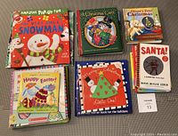 Seven toddler holiday-themed books arranged on carpet, titles include snowman, Christmas carol, Easter and Christmas pop-up books, and a lift-the-flap Santa book.