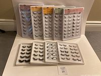 11 trays of false lashes arranged on a white surface. Various styles visible with labels on trays.