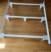 Photo showing white queen bed frame assembled on floor with wooden slats and legs visible, condition excellent.