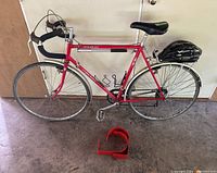 Full side view of red Maruishi 10 speed road bike with black helmet on rear rack and red bike rack brackets on ground.