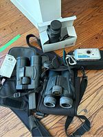 Nikon and EXP binoculars, Pentax digital camera, and uni-nocular monocular displayed on a wooden floor