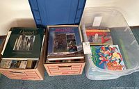 Three plastic and cardboard boxes containing a variety of hardcover and softcover books including children's books, with visible titles 'Queen's The First One Hundred & Fifty Years' and 'Britain and Ireland'.