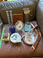 Photo of assorted decorative wares on couch including glass candlesticks, wooden clock, glass pitcher, blue glass sphere, agate slice, ceramic plates, painted boomerang, wooden mallet, floral ceramic box, and ashtray.
