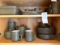 Wide view of wooden cabinet shelf with Denby stoneware, mugs, teapot, coffee pot, stacked plates and bowls in olive green textured pattern.