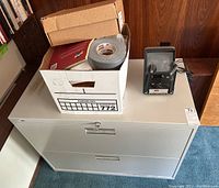 Gray metal lateral file cabinet with two drawers and lock, cardboard box of office supplies and duct tape on top, 8mm film editor machine on top right corner of cabinet