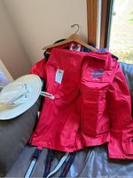 Photo of a red Mariner wet skins rain jacket with multiple front pockets and adjustable straps, hanging on a hanger next to an off-white wide-brimmed Tilly hat.