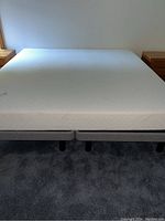 White king size mattress on two grey bed frames on carpeted floor with two wooden nightstands in the background.