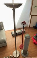 Tall metallic torchiere floor lamp, red floor lamp, silver tower fan on wood floor near couch and window