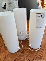 Three white frosted glass cylindrical lamps in upright position on wood floor, power cords visible.