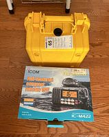 Yellow waterproof hard carrying case and Icom IC-M422 marine VHF radio box placed on wooden floor.