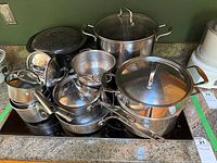 Set of various stainless steel pots and pans with lids placed on a countertop including a speckled enamel pot
