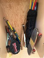 Photo showing various rackets including tennis rackets with covers, wooden and metal baseball bats, grass hockey sticks, a canister of tennis balls, and partial view of a skateboard leaning against a wooden wall.