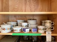 Full shelf view displaying stacked Wedgwood dinner plates, multiple mugs, lidded sugar bowl, and teapot with Quince pattern.