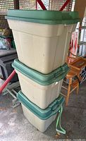 Stack of three beige plastic storage totes with green lids, outdoors on a concrete surface.
