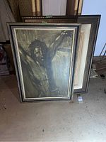Full view of the framed artwork depicting a crucified figure in bas-relief or charcoal style, leaning against a wall on a basement floor.