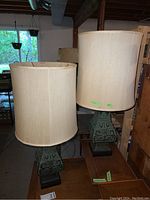 Photo showing two lamps with Asian style green patinated metal bases and tall beige shades that have staining. Lamps are on a wooden surface in a basement setting.