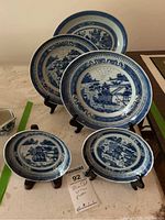 Five blue and white porcelain plates arranged with wooden stands, showing the Blue Willow pattern landscape scenes.