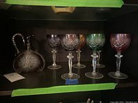 Photo of 6 colored cut to clear wine glasses and one matching pitcher showing shape, size, and color variety in a dark display setting.