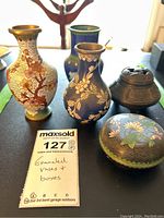 Five pieces including four small enamel vases and two decorative boxes displayed on table. Close detail of floral enamel work and metal box.