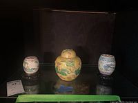 Three ginger jars displayed on a glass shelf including a yellow lidded jar and two smaller white jars on black stands.