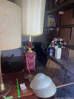 Red glass base lamp with cream fabric shade and decorative gold trim seen on a wooden table in basement setting.