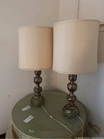 Two brass-tone table lamps with ivory fabric shades on round green table