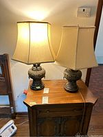 Photo of two lit table lamps with Asian style urn-shaped bases and cream fabric square shades on a wooden cabinet.