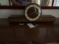 Front view of vintage mantle clock sitting on table, showing wood case, round clock face with gold-tone numbers and included winding key in front.