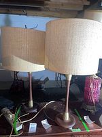 Pair of mid century modern wooden lamps with beige woven fabric drum shades placed on a table in a basement with rodent damage evidence.