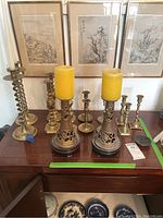 Photo showing two large decorative brass holders with yellow pillar candles centered on a wood table with framed artwork behind