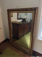Full view of large rectangular mirror with gold ornate frame leaning against wall, showing part of room reflection.