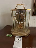 Full view of quartz clock showing gold-tone metal frame, glass panels, Roman numerals, and hexagonal base with some staining.