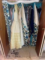 Closet with assorted vintage dresses and garments covered by floral protective garment bags, including a visible pale yellow formal dress with lace and embroidery detail.